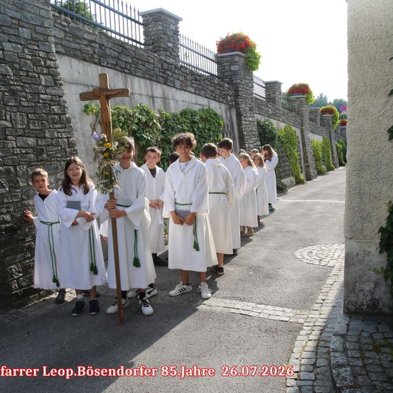 Eine Gruppe von Kindern in weißen Roben, die ein Kreuz und Blumen halten, steht in einer Reihe auf einer Straße.