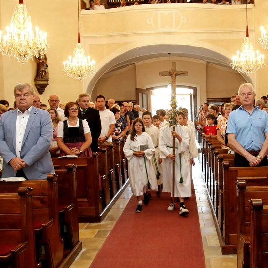 Eine Gruppe von Menschen, einschließlich Kinder, gehen den Gang einer Kirche entlang, halten ein Kreuz und einen Stab.