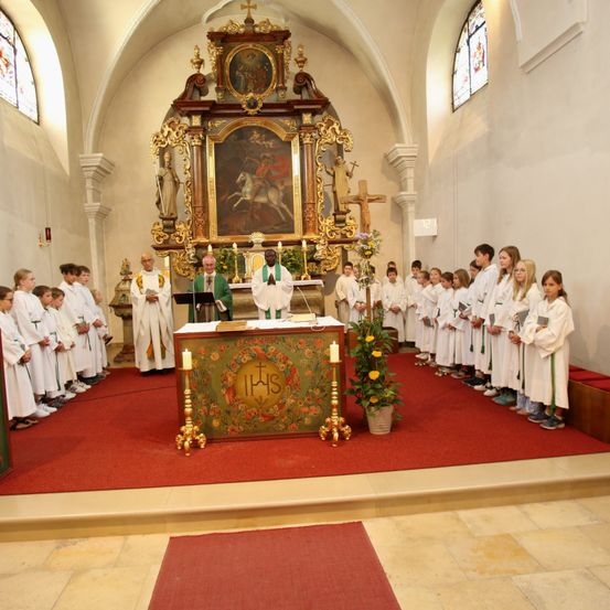 Ein Priester steht an einem Altar in einer Kirche mit einem Chor von Kindern in weißen Gewändern. Der Altar ist mit Blumen und Kerzen geschmückt.