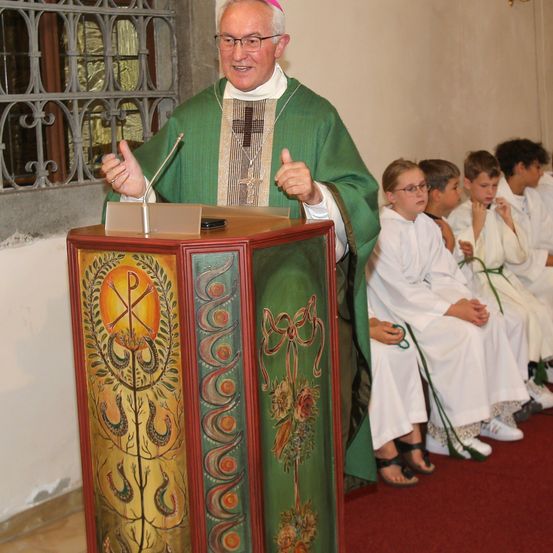 Ein Priester in einer grünen Robe steht an einem Podium und hält eine Rede mit einem Mikrofon. Hinter ihm sitzen mehrere Kinder in weißen Gewändern auf einem roten Teppich.