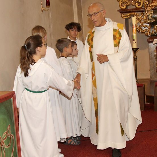 Ein Priester in einer weißen Robe mit einem grünen Gürtel gibt einem Mädchen in einem weißen Kleid die Hand in einer Kirche.
