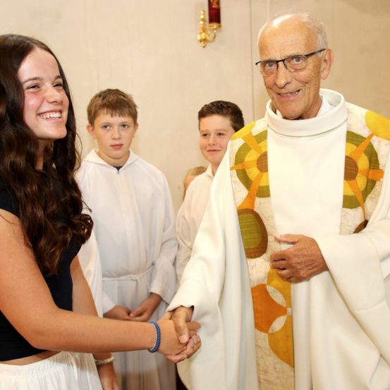 Ein Priester in einem farbenfrohen Gewand schüttelt die Hand einer lächelnden Mädchen, mit zwei Jungen in weißen Gewändern hinter ihnen.