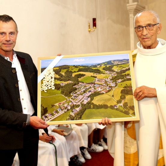 Ein Priester und ein Mann in Anzug halten ein gerahmtes Gemälde einer Dorflandschaft.