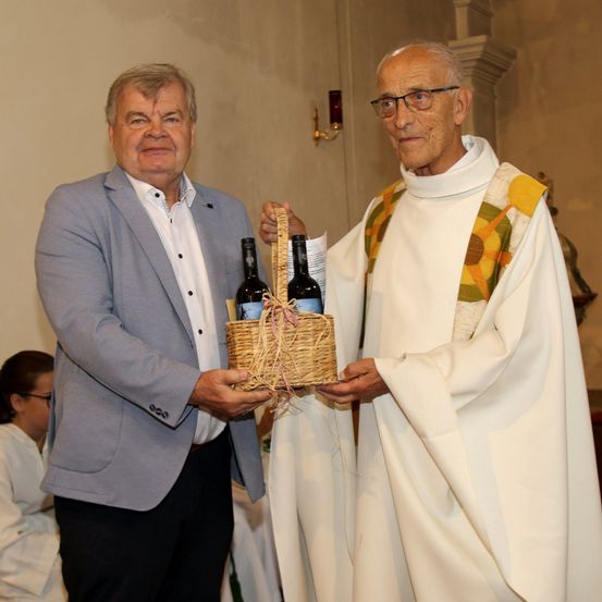 Zwei Männer, einer in einem grauen Anzug und der andere in einer weißen Robe, halten einen Korb mit zwei Flaschen Wein. Sie stehen vor einer Wand mit einem Kerzenhalter.