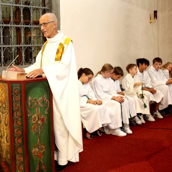 Ein älterer Priester steht an einem Podium und spricht zu einer Gruppe von Kindern in weißen Gewändern, die auf einem roten Teppich sitzen.
