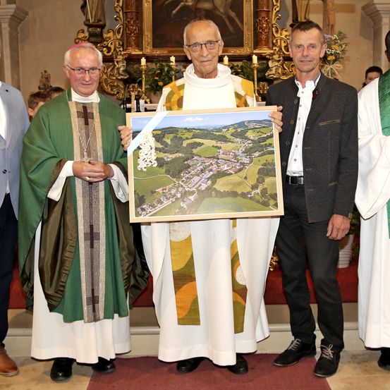 Drei Männer stehen in einer Kirche, einer hält eine gerahmte Luftaufnahme eines Dorfes. Der Mann auf der linken Seite trägt eine grün-weiße Robe, der Mann in der Mitte trägt eine weiß-gelbe Robe und der Mann auf der rechten Seite trägt einen dunklen Anzug.