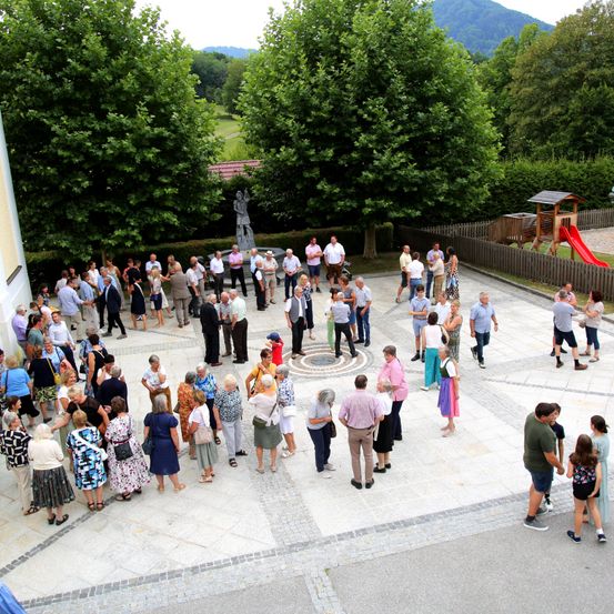 Eine Gruppe von Menschen versammelt sich in einem Außenbereich, möglicherweise für eine Veranstaltung. Sie stehen im Kreis um eine Statue. Einige tragen Hüte und Schuhe, andere haben Taschen dabei. In der Umgebung gibt es Bäume und einen Spielplatz. In der Ferne befindet sich ein Haus mit einem roten Dach.