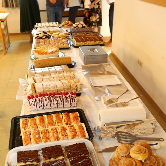 Ein langer Tisch ist mit verschiedenen Desserts wie Kuchen, Gebäck und Zimtschnecken gefüllt. Im Hintergrund stehen Personen.