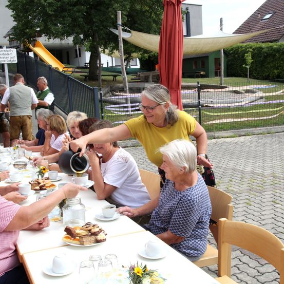 Eine Gruppe älterer Frauen sitzt um einen Tisch herum und genießt eine Mahlzeit und Getränke. Eine Frau in einem gelben Shirt schenkt einer anderen Frau Kaffee ein. Im Hintergrund befindet sich ein Spielplatz mit einer Rutsche und einem Zaun.