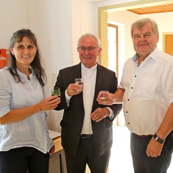 Drei Personen, zwei Männer und eine Frau, stehen zusammen, lächeln und halten Gläser mit Wasser. Die Frau trägt ein hellblaues Oberteil, und die Männer sind in Anzügen gekleidet. Der Hintergrund zeigt eine weiße Wand und eine Tür.