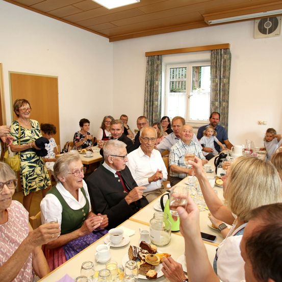Eine Gruppe älterer Menschen sitzt an einem langen Tisch in einem Raum, einige klatschen und lächeln, während andere essen und trinken. Der Raum hat weiße Wände, eine Holzdecke und ein Fenster mit Vorhängen. Auf dem Tisch stehen Teller, Tassen und andere Gegenstände.