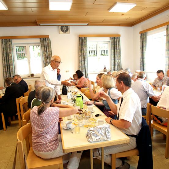 Eine Gruppe älterer Menschen sitzt an einem langen Tisch in einem Speisesaal und isst. Ein Mann in einem weißen Hemd steht und spricht mit ihnen.