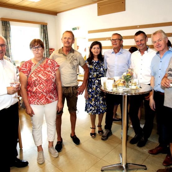 Eine Gruppe von Erwachsenen, sowohl Männer als auch Frauen, stehen um einen kleinen Tisch mit Getränken und Snacks und lächeln für ein Foto in einem Raum.