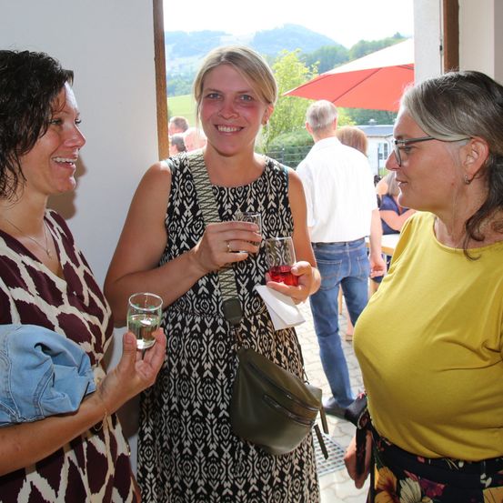 Drei Frauen stehen in einer Türöffnung, lächeln und halten Getränke. Eine Frau trägt ein gemustertes Kleid und eine Tasche, während eine andere ein Glas Wasser hält. Im Hintergrund steht ein Mann in einem weißen Hemd in der Nähe eines roten Schirms.