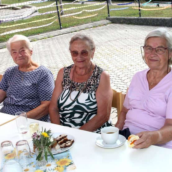 Drei ältere Frauen sitzen an einem Tisch mit einem Teller Essen, einer Tasse Kaffee und einem Glas Wein. Sie lächeln und scheinen ihre Mahlzeit zu genießen.