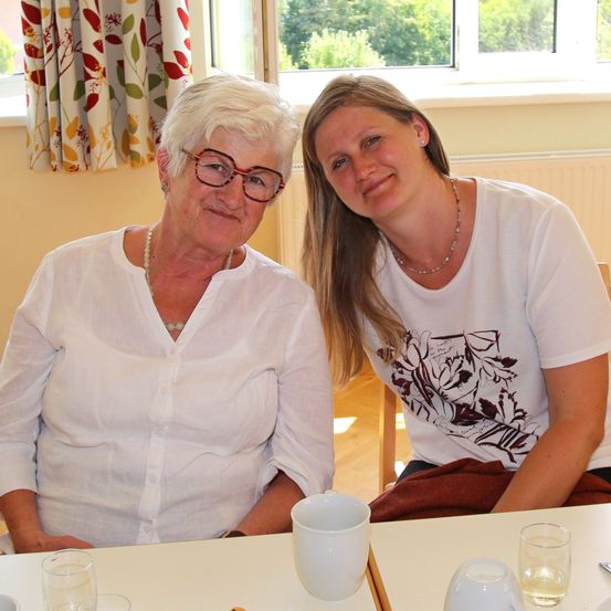 Zwei Frauen sitzen an einem Tisch, eine mit Brille und Halskette, die andere trägt ein bedrucktes Shirt. Sie lächeln und posieren für ein Foto. Hinter ihnen ist ein Fenster mit Blick auf Bäume.