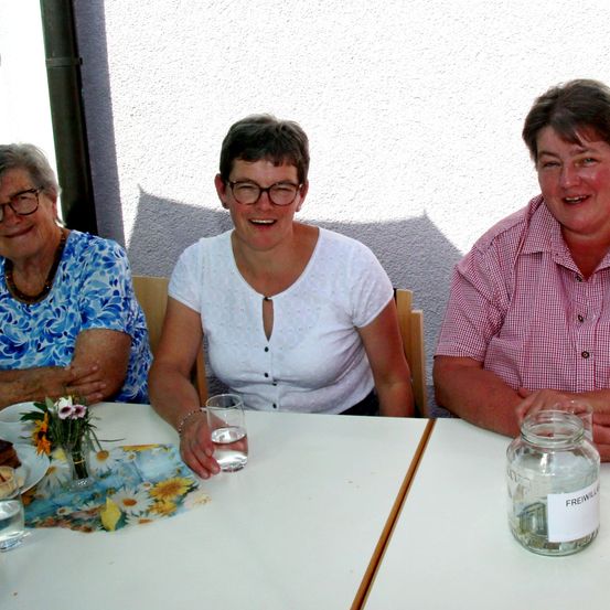 Drei Frauen sitzen an einem Tisch und lächeln. Die Frau links trägt ein Blumenhemd, die mittlere eine weiße Bluse und die rechte ein rosa kariertes Hemd. Auf dem Tisch steht ein Glas mit der Aufschrift 'Freiwillig'.