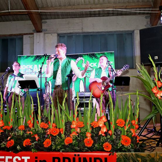 Eine Band tritt auf der Bühne auf, mit einem Banner hinter ihnen, das 'JOCK AIR' besagt. Der Leadsänger hält ein Mikrofon, während ein anderes Mitglied die Gitarre spielt. Die Bühne ist mit orangefarbenen Blumen und grünen Pflanzen geschmückt.