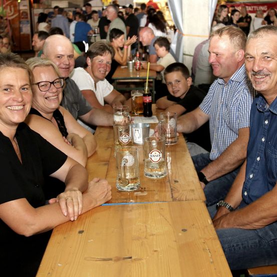 Eine Gruppe von Menschen sitzt an einem Tisch in einem überfüllten Biergarten, lächelt und posiert für ein Foto. Leere Gläser und eine Coca-Cola-Flasche stehen auf dem Tisch.