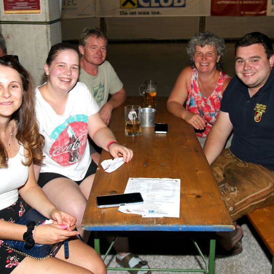 Fünf Personen sitzen um einen Holztisch und lächeln für ein Foto. Zwei Frauen und drei Männer, mit Getränken und einer Speisekarte auf dem Tisch.