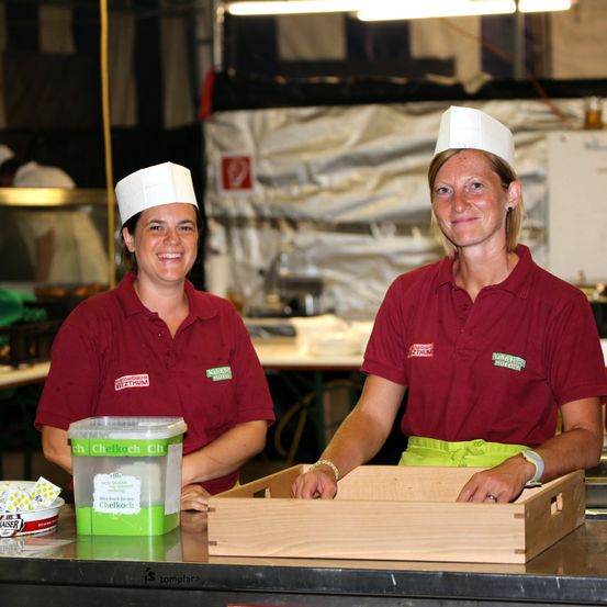 Zwei Frauen in roten Uniformen und weißen Kochmützen stehen hinter einer Theke in einer Küche, eine hält einen Behälter und die andere eine Holzschale.