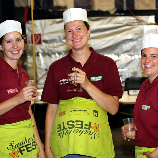 Drei Frauen in Kochmützen und Schürzen, lächelnd und halten Getränke in einer Fabrikumgebung. Die Frau in der Mitte hält ein Glas Bier.