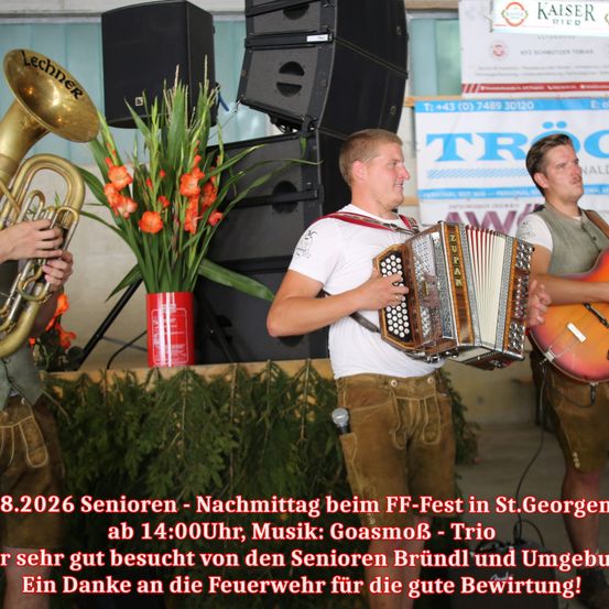 Drei Männer spielen auf der Bühne mit Musikinstrumenten. Der Mann in der Mitte spielt Akkordeon, während die anderen Trompete und Gitarre spielen. Die Bühne ist mit einer Pflanze und einem Feuerlöscher dekoriert. Das Bild hat einen Wasserzeichen mit Text auf Deutsch.
