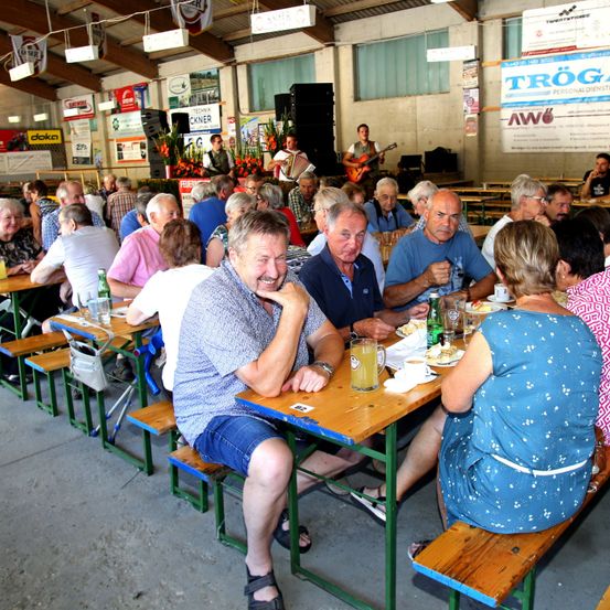 Eine Gruppe von Menschen sitzt an Tischen in einem großen Innenbereich, möglicherweise einem Restaurant. Ein Mann spielt Gitarre, während ein anderer Akkordeon spielt. Auf den Tischen stehen Flaschen, Tassen und Teller.