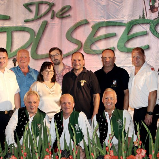 Eine Gruppe Erwachsener in traditioneller Kleidung posiert für ein Foto vor einem Banner mit grünem Text.