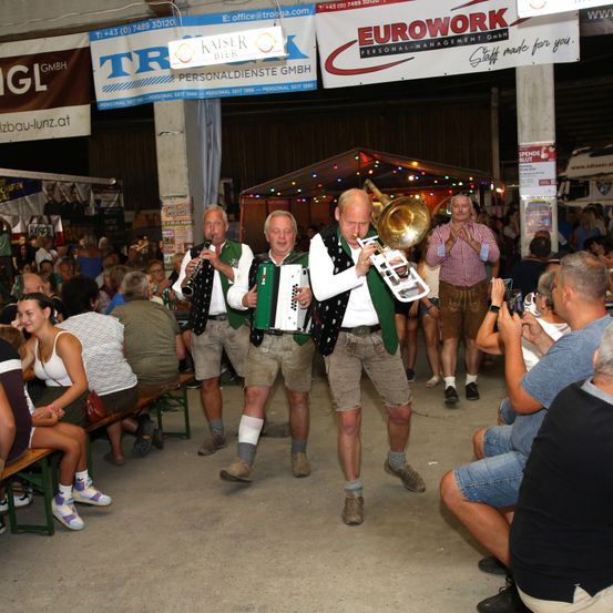 Drei Männer in traditioneller bayerischer Kleidung spielen Musikinstrumente bei einer lebhaften Veranstaltung, umgeben von einer Menge von Zuschauern.