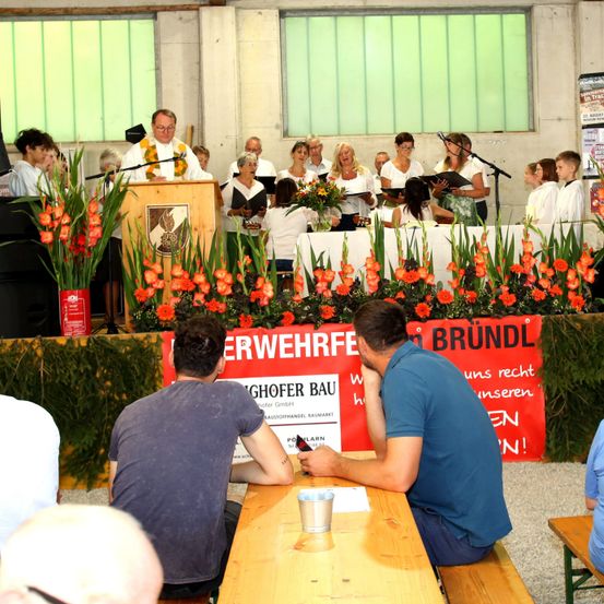 Ein Chor singt auf der Bühne mit einem Banner, das 'Brundl' im Hintergrund zeigt, während zwei Männer am Tisch im Vordergrund sitzen.