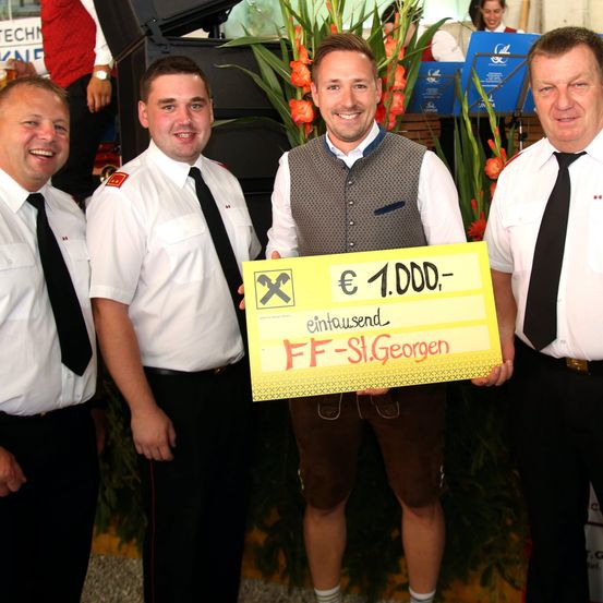 Vier Männer in Uniform stehen zusammen, einer hält einen gelben Scheck über 1.000 Euro, mit einem lächelnden Gesichtsausdruck.