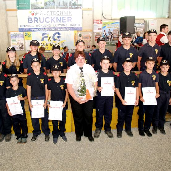 Eine Gruppe junger Personen in Uniform, die Urkunden halten, steht in einer Reihe mit einer Frau in der Mitte. Hinter ihnen ist ein Banner mit dem Text 'Haustechnik Bruckner' angebracht.