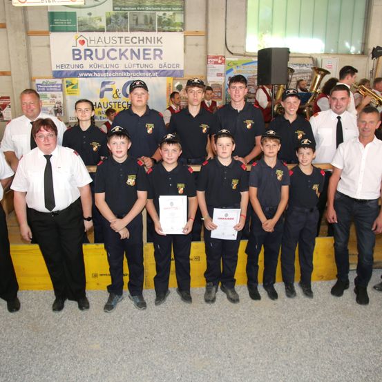 Eine Gruppe junger Jungen in Uniform mit Urkunden steht vor einem Banner mit Logo und Text. Sie werden von Erwachsenen umgeben, einige halten Musikinstrumente.