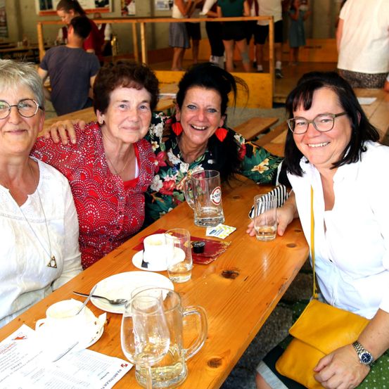 Vier Frauen sitzen an einem Holztisch, lächeln und posieren für ein Foto. Zwei von ihnen tragen eine Brille, und eine hält ein Glas Bier. Der Tisch hat Teller, Tassen und eine Speisekarte. Im Hintergrund stehen und sitzen Leute auf Bänken.