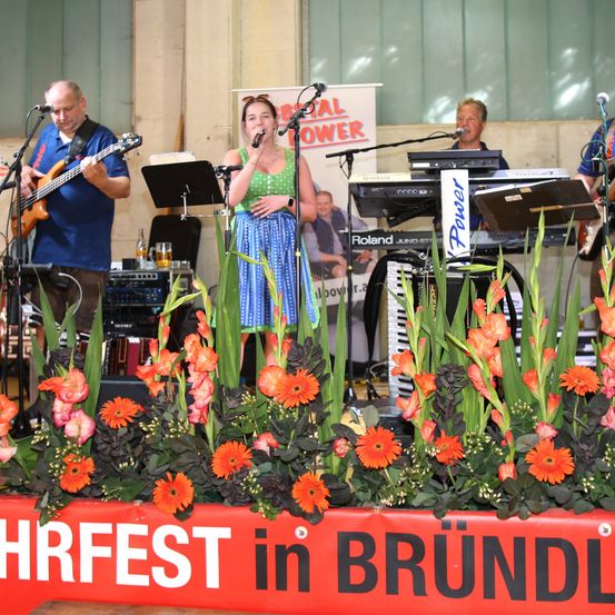 Eine Band tritt bei dem 'Hirfest in Brundl' Festival auf der Bühne auf, mit einer Sängerin, einem Gitarristen und einem Keyboarder. Die Bühne ist mit orangefarbenen Blumen geschmückt.