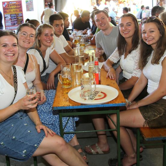 Eine Gruppe von Menschen sitzt an einem Tisch in einer Bar, lächelt und hält Gläser mit Bier in der Hand. Sie sind von anderen Gästen umgeben, und auf dem Tisch steht ein Teller mit dem Wort 'Kaiser' darauf.
