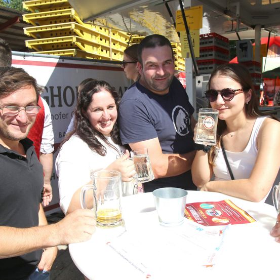 Vier Personen lächeln und posieren für ein Foto mit Bierkrügen bei einer Outdoor-Veranstaltung. Dahinter ist ein Stapel gelber Kisten zu sehen.