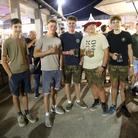 Fünf junge Männer in lässiger Kleidung und traditioneller bayerischer Tracht stehen zusammen und halten Gläser Bier bei einer nächtlichen Veranstaltung im Freien.