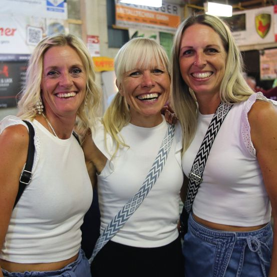 Drei Frauen mit blonden Haaren, die lächelnd für ein Foto posieren. Sie tragen weiße Oberteile und stehen eng beieinander.