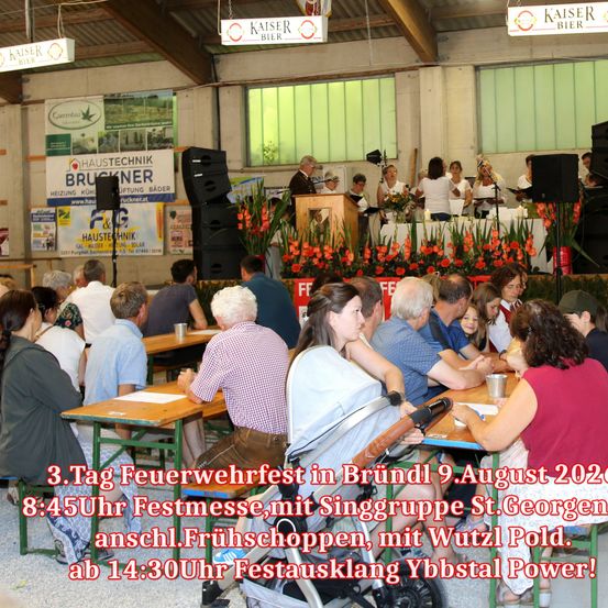 Eine Menschenmenge sitzt an Tischen in einem Saal. Eine Gruppe von Menschen tritt auf der Bühne mit Blumen und einem Podium auf. Der Text auf dem Banner lautet '3. Tag Feuerwehrfest in Bründl 9. August 2020'.