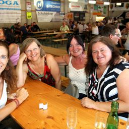 Vier Frauen sitzen an einem Tisch in einem überfüllten Biergarten und lächeln für die Kamera. Hinter ihnen sitzen weitere Personen an Tischen und Bänken, wobei Banner und Schilder an den Wänden sichtbar sind.