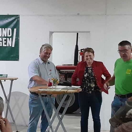 Drei Personen stehen um einen Tisch mit einer Flasche und Papieren. Ein Mann in einem weißen Hemd lächelt, eine Frau in einer roten Jacke steht neben ihm und ein Mann in einem grünen Hemd steht hinter ihnen. Ein Feuerlöscher ist an der Wand hinter ihnen montiert.