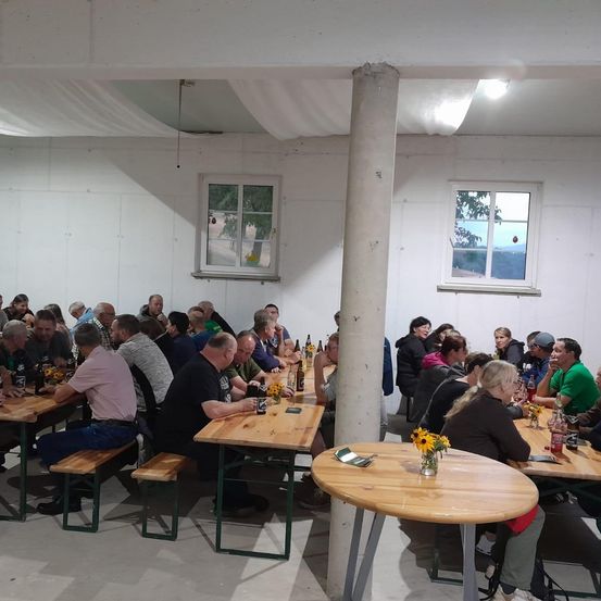 Eine Gruppe von Menschen sitzt um Tische herum in einem Raum mit weißen Wänden, Holztischen und Fenstern. Einige tragen Hüte und Brillen, und es gibt Flaschen und Blumen auf den Tischen.