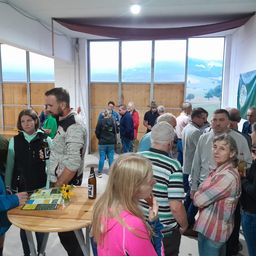 Eine Gruppe von Menschen versammelt sich in einem Raum mit großen Fenstern, die eine malerische Landschaft überblicken. Einige halten Getränke in der Hand, und ein Tisch im Vordergrund hat eine Karte und eine Flasche.