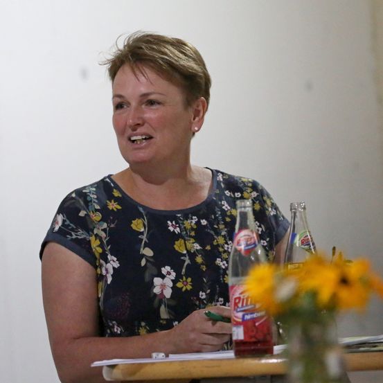 Eine Frau mit kurzen blonden Haaren sitzt an einem Tisch, lächelt und hält einen Stift. Sie trägt ein Blumenhemd und hat eine Flasche Limonade und einen Blumenvase auf dem Tisch.