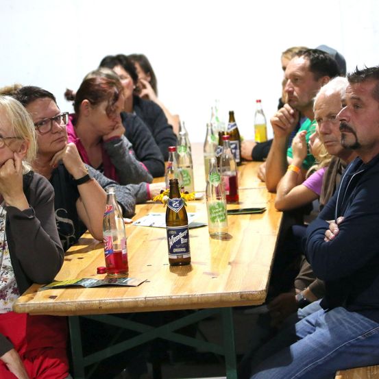 Eine Gruppe von Menschen sitzt um einen Holztisch mit verschiedenen Flaschen und einer Speisekarte, einige tragen Brillen und Armbänder, einer mit verschränkten Armen.