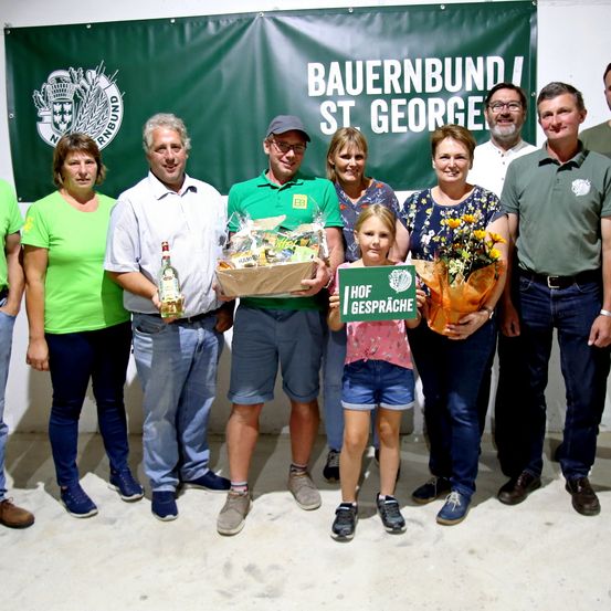 Eine Gruppe von Menschen in grünen Shirts und Hüten posiert für ein Foto mit einem Banner, das Bauernbund St. George liest. Ein Mann hält eine Flasche und einen Korb, während ein Mädchen ein Schild hält, das Hof Gespraeche liest.