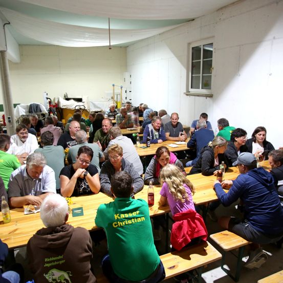 Eine große Gruppe von Menschen sitzt an langen Holztischen in einem Raum, einige unterhalten sich, während andere essen. Der Raum hat weiße Wände und ein Fenster. Ein Mann in einem grünen Shirt mit dem Wort 'Kasser Christian' darauf sitzt an einem Tisch.