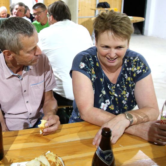 Ein Mann und eine Frau sitzen an einem Tisch, der Mann isst ein Sandwich und die Frau lächelt. Hinter ihnen sitzen und stehen mehrere Personen in einem Raum.